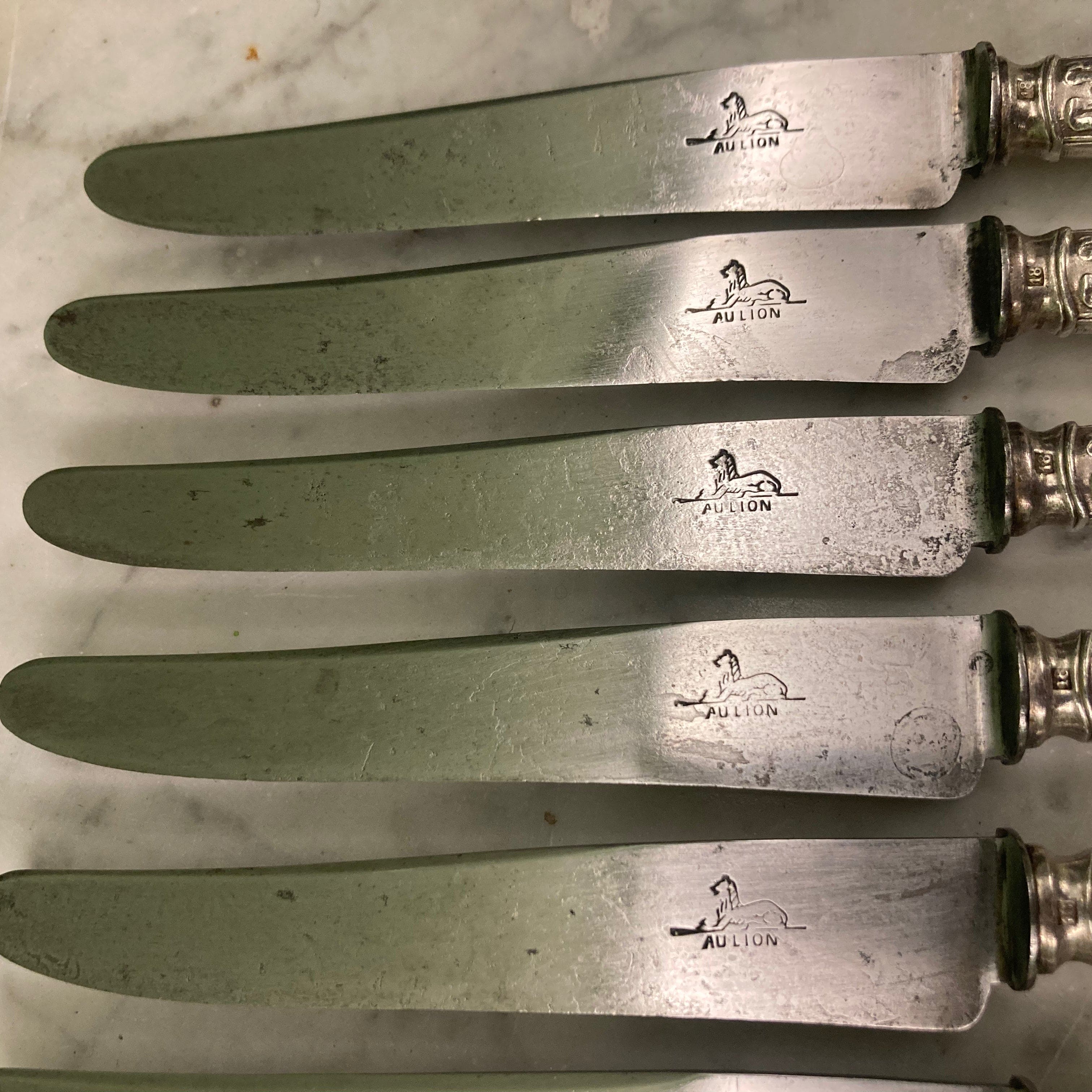 12 couteaux à entremets en métal argenté et lames acier. Modèle rubans croisés. Marque: Au Lion