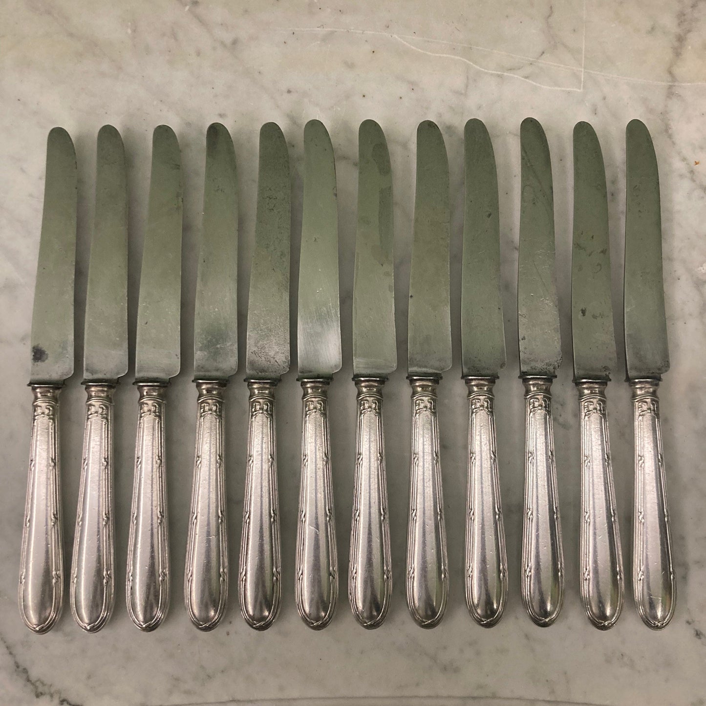 12 couteaux à entremets en métal argenté et lames acier. Modèle rubans croisés. Marque: Au Lion