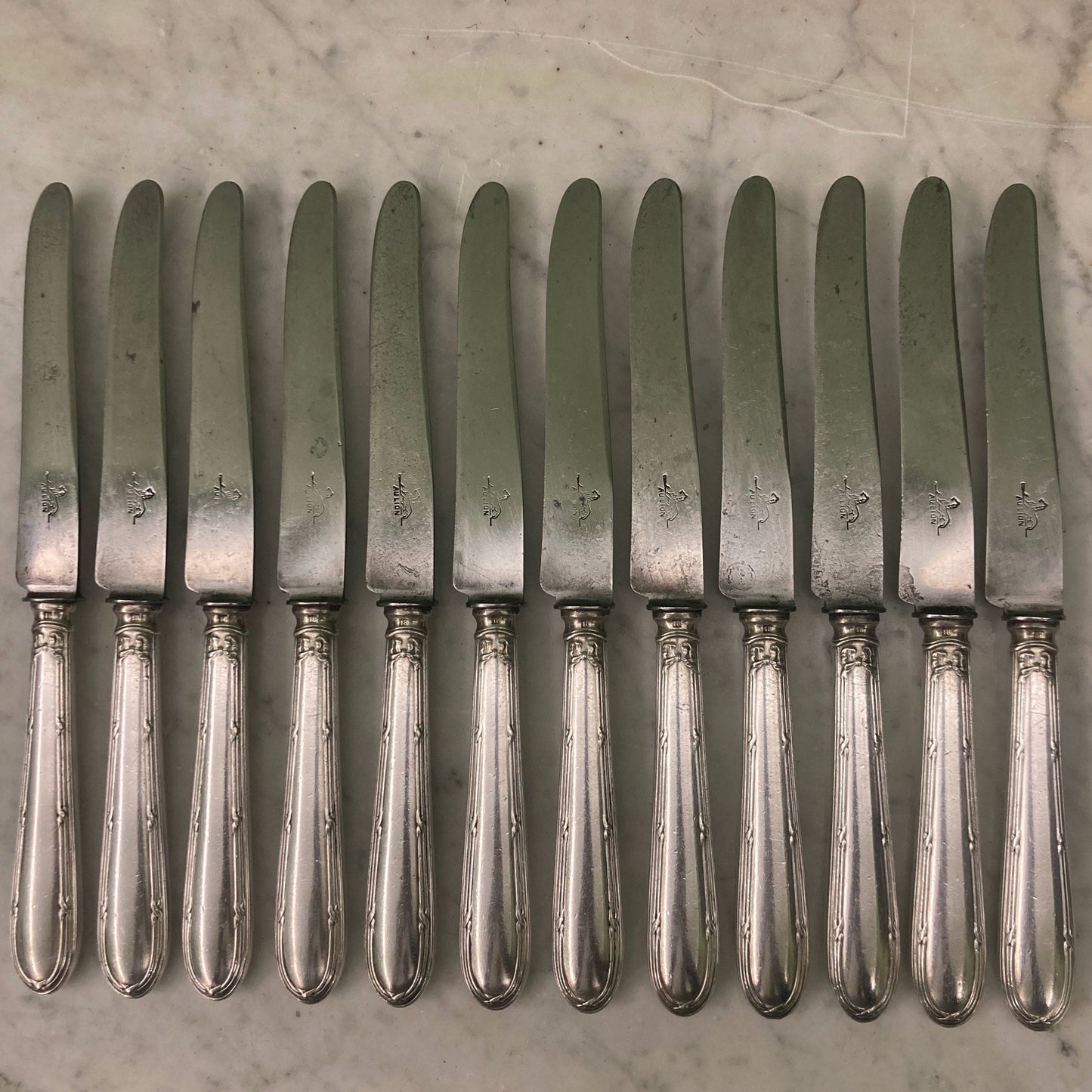 12 couteaux à entremets en métal argenté et lames acier. Modèle rubans croisés. Marque: Au Lion